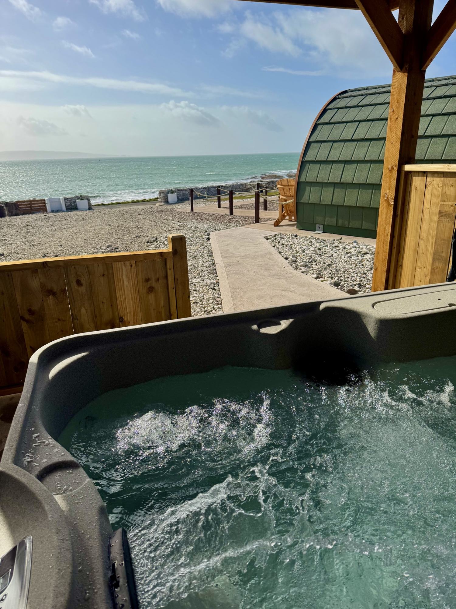 The hot tub at Pod One bubbling in daylight, with the green shingled pod and the turquoise sea behind.